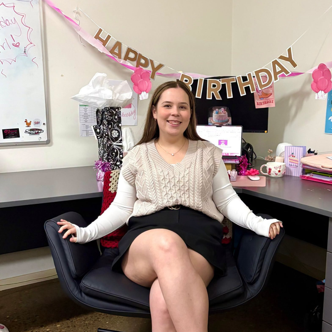 Woman seated in a modern office chair at a desk, smiling, with a pink birthday banner reading 'HAPPY BIRTHDAY' behind her.